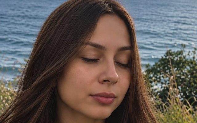 Mulher em momento de presença e quietude, de olhos fechados, com o mar ao fundo